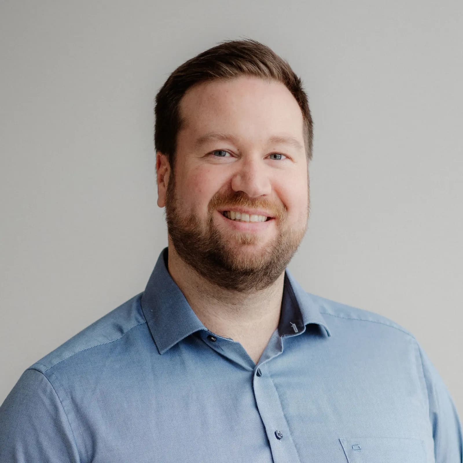 Birk Kulwicki steht lächelnd vor einer grauen Wand. Er trägt ein blaues Hemd.